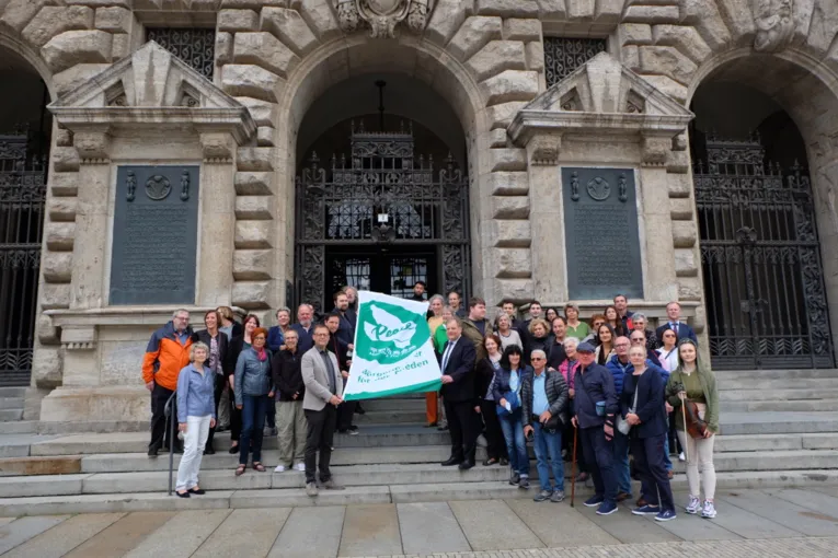 Eine gro&szlig;e Gruppe von Menschen unterschiedlichen Geschlechts und Alters h&auml;lt die gr&uuml;n-wei&szlig;e Mayors-for-Peace-Flagge vor dem Rathaus in Leipzig in die Kamera.